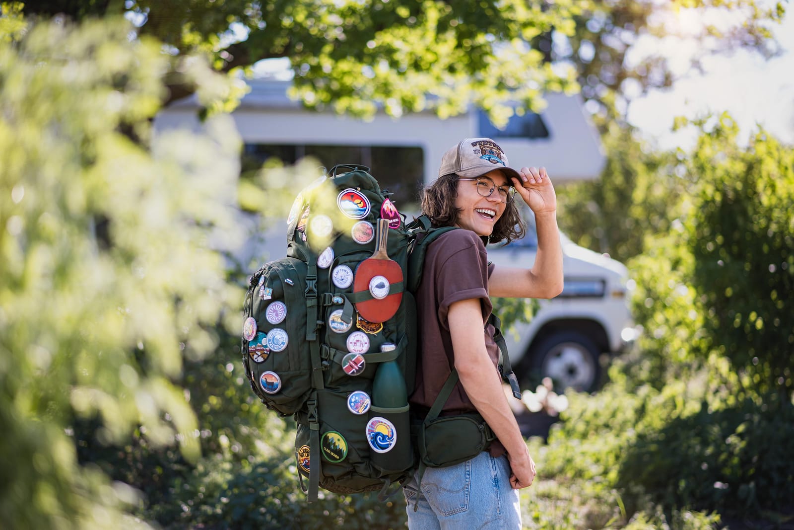 Eine lachende Person mit Kappe und einem Rucksack voller Aufnäher steht zwischen grünen Büschen.